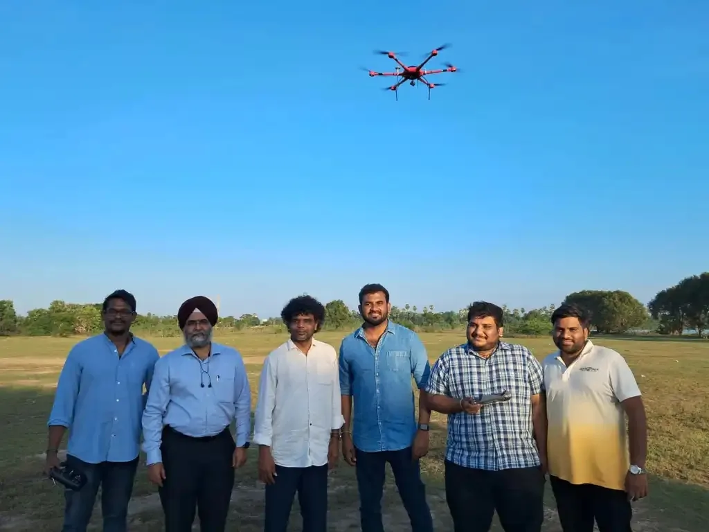 Team of six posing on a grass field while a red hexacopter drone hovers overhead during a test flight